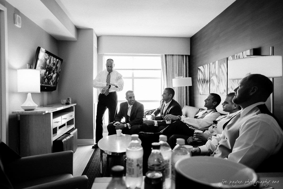A black and white photo of six men dressed in suits sitting in a hotel room, watching TV. One man is standing and adjusting his tie while others are seated, engaging in conversation. Bottles of water are visible on the table in the foreground.