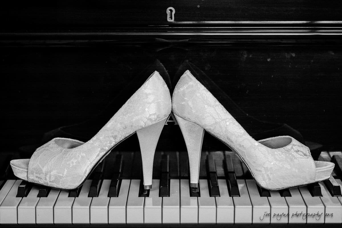 A black and white image of two high-heeled shoes positioned back-to-back on piano keys. The shoes have a lace texture, and the piano is partially visible in the background.