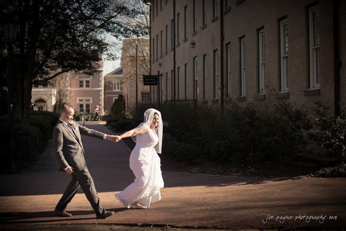 A bride and groom joyfully walk hand in hand along a sunlit brick pathway between two buildings. The bride, in a white gown and veil, looks back at the groom, who is in a gray suit, as they move towards the sunlight.