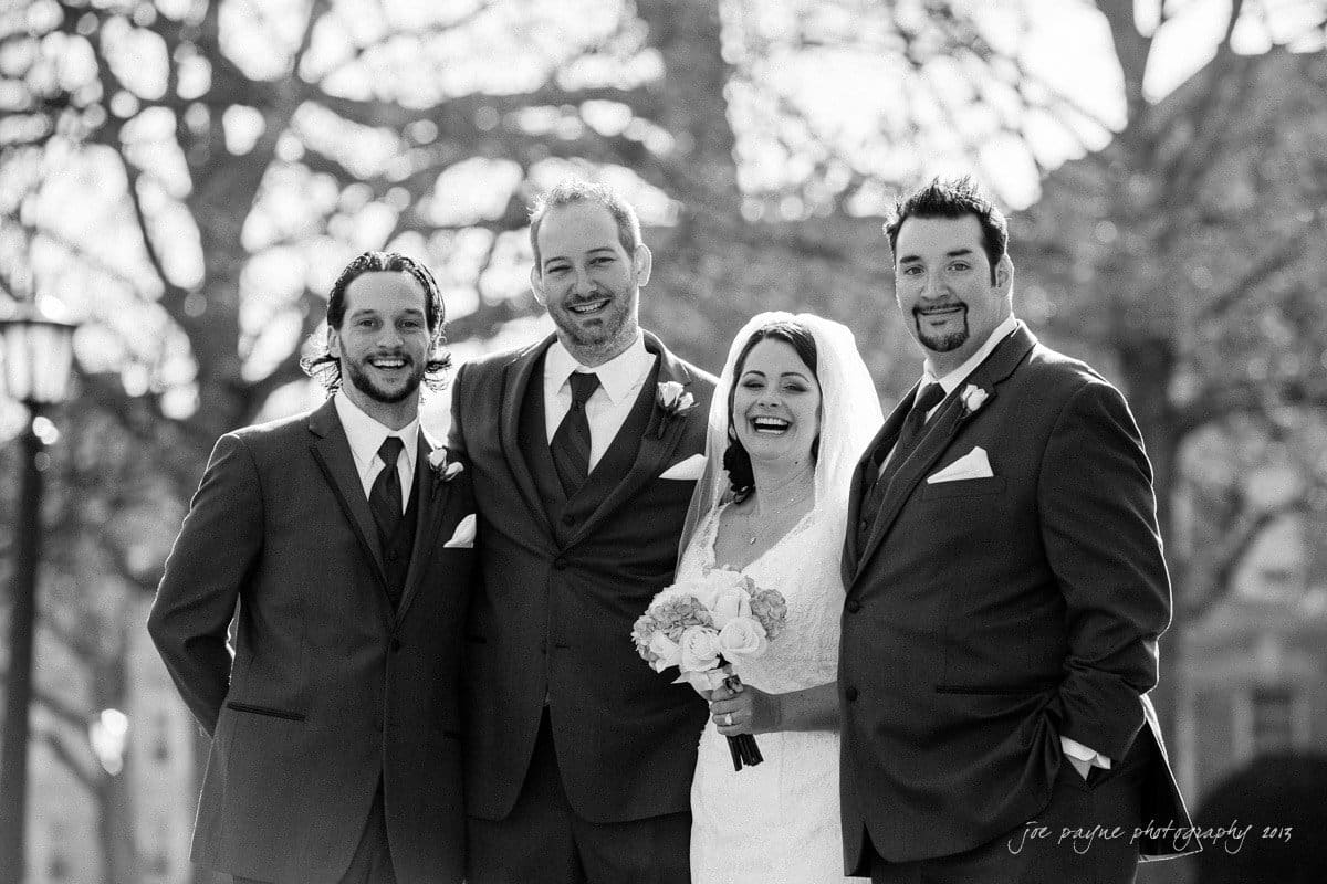 A black and white photo of a wedding group. A bride in a white gown and veil holds a bouquet, standing with three men in suits. They are smiling and posed closely together outdoors, with soft focus trees in the background.