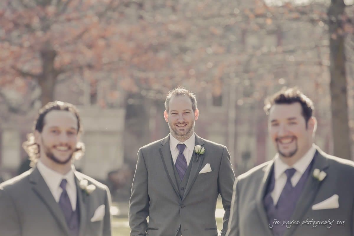 Three men in suits standing outdoors, surrounded by trees. The man in the center is in focus, while the two men on either side are softly blurred. They are dressed in gray suits with purple ties and boutonnieres, suggesting a formal occasion.