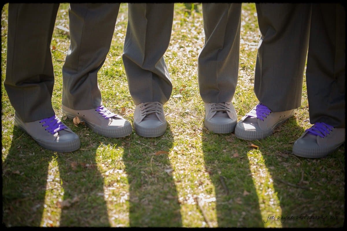 Four people wearing gray shoes stand on grass. Three pairs have purple laces, one has gray. They are wearing gray pants, and long shadows are cast on the ground by the sunlight.