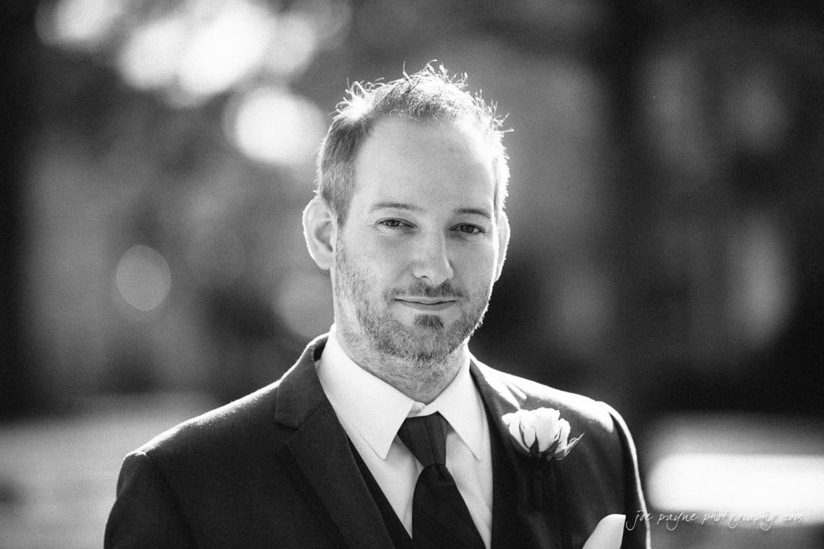 A black and white portrait of a man in a suit and tie, standing outdoors. He has a slight smile and a boutonniere on his jacket. The background is softly blurred.