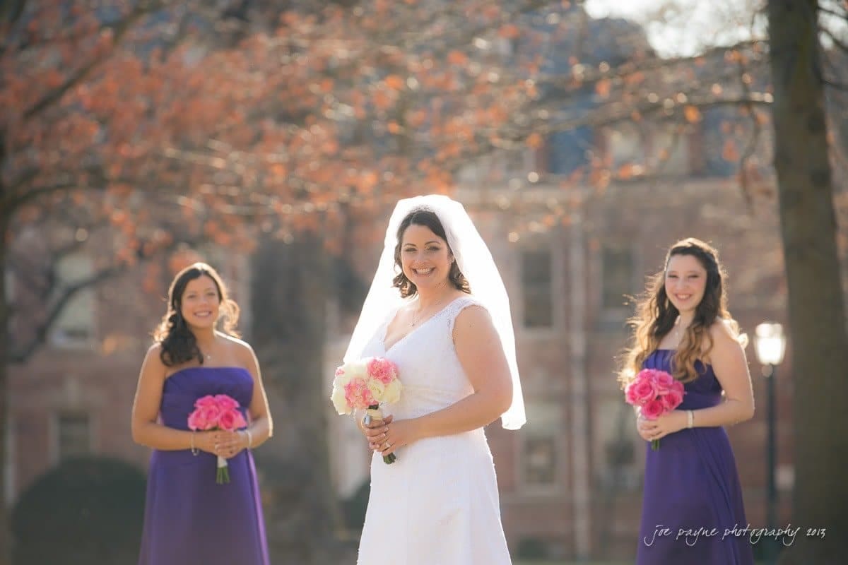 A bride in a white dress and veil stands smiling outdoors, holding a bouquet of pink and white flowers. Two bridesmaids in purple dresses stand on either side, each holding bouquets of pink flowers. They are in a sunny setting with trees and a building.