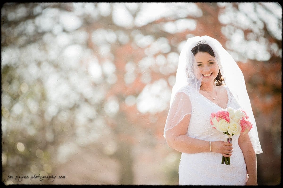 A bride in a white dress and veil smiles while holding a bouquet of pink and white flowers. She stands outdoors with soft focus trees in the background.