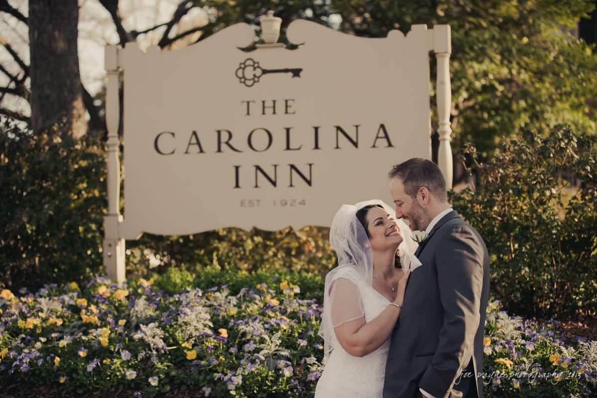 A couple in wedding attire smiles at each other in front of a sign for The Carolina Inn, established in 1924. They are surrounded by green foliage and colorful flowers.