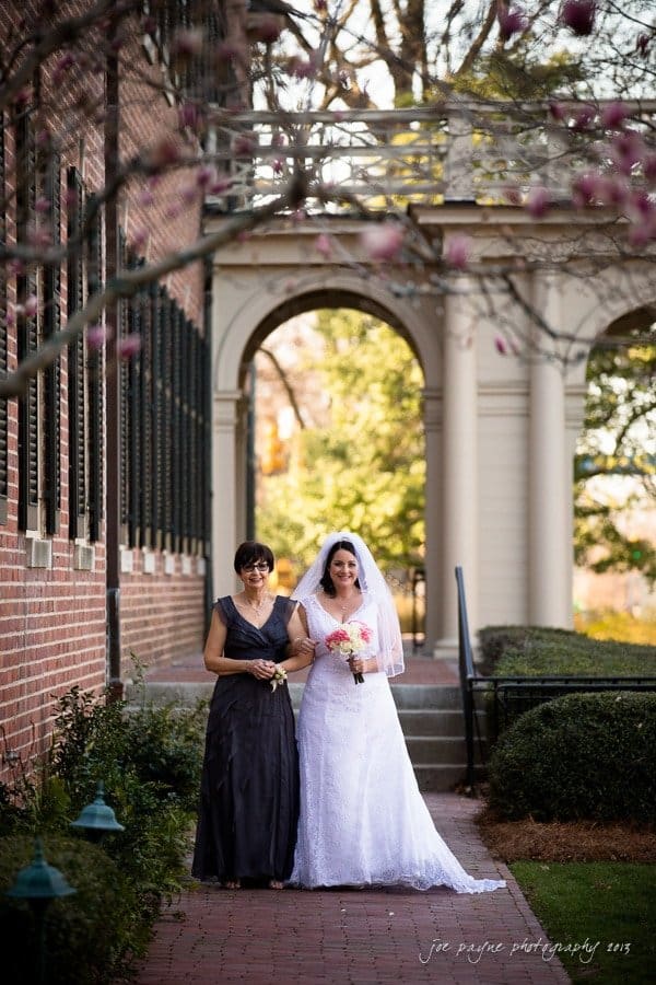 A bride in a white gown and veil walks arm in arm with a woman in a dark dress along a brick pathway. They are surrounded by spring blossoms and trees, with columns in the background.