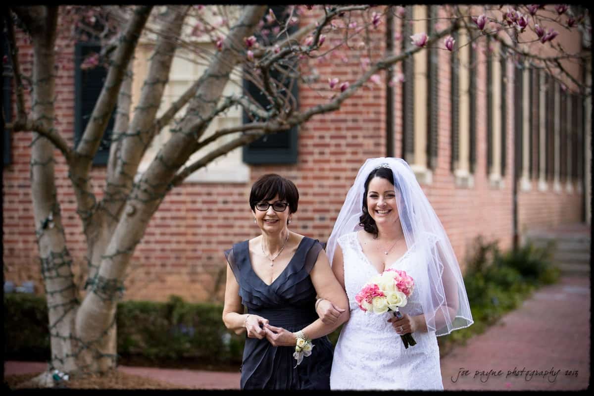 A bride in a white dress and veil walks arm in arm with a woman in a dark dress. They smile as they stroll down a path lined with a brick building and blooming tree. The bride holds a bouquet of white and pink flowers.