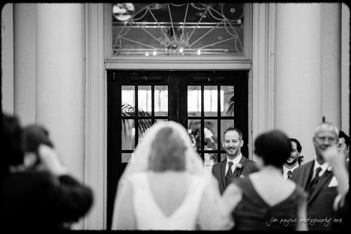 A bride in a wedding dress walks toward a groom in a suit, who is smiling. They are flanked by guests on either side, capturing a joyful moment at a wedding ceremony.