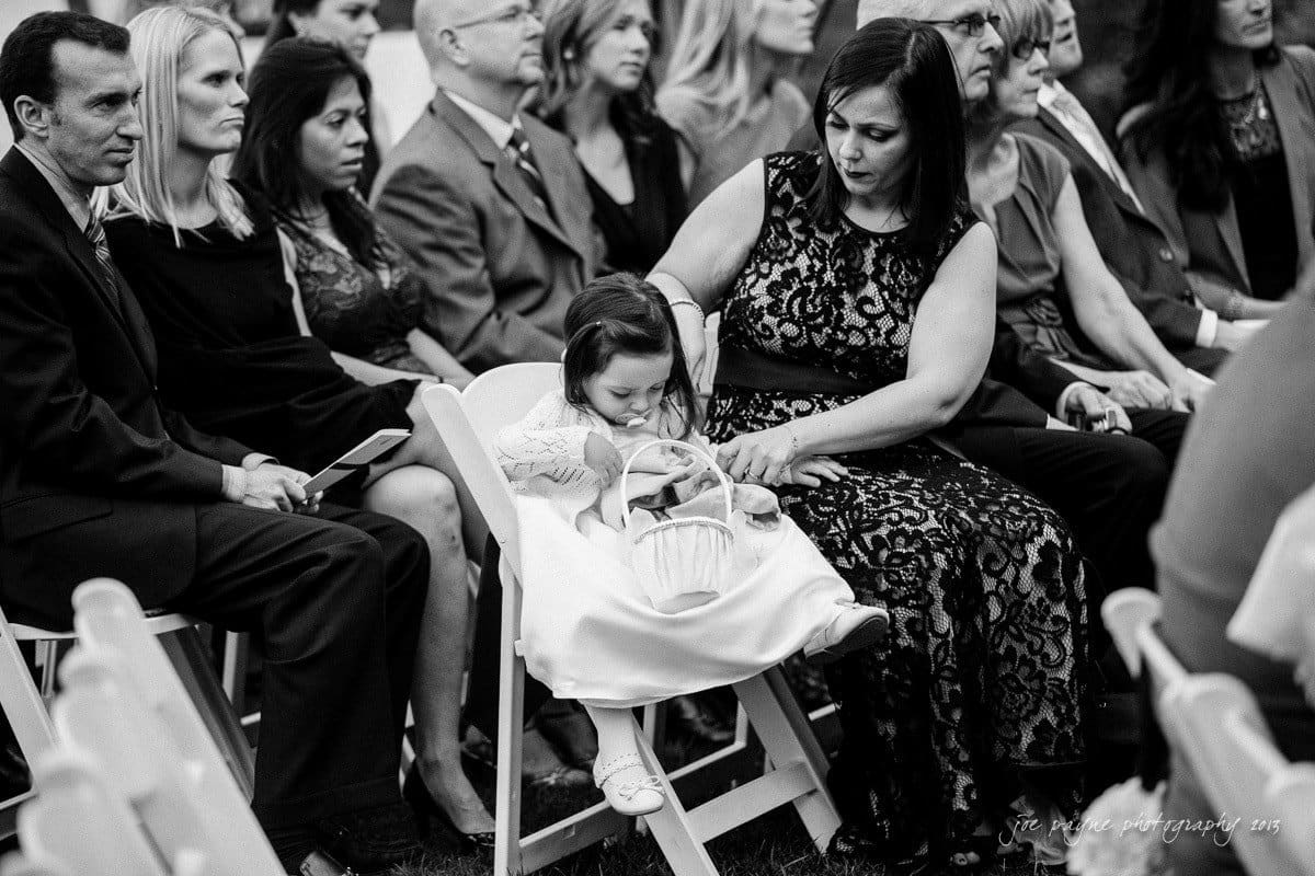 A black and white photo of a seated group at an outdoor event. A child in a white dress leans on a woman wearing a lace dress, holding a basket. Other attendees, dressed formally, sit attentively in the background.