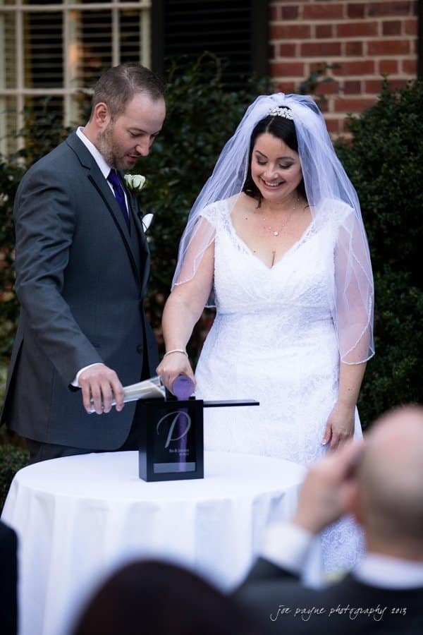 A bride and groom participate in a unity ceremony by pouring two different-colored sands into a single container during their outdoor wedding. The bride wears a white gown and veil, while the groom is in a gray suit. They are smiling and joyful.