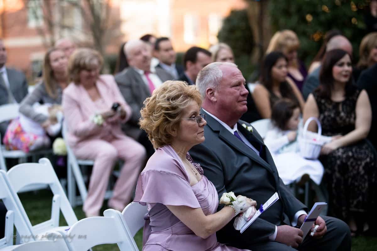 An outdoor wedding ceremony with guests seated. A woman and a man in formal attire are sitting in the foreground, focused on the event. Other attendees, including children, are visible in the background. White chairs and greenery surround the scene.