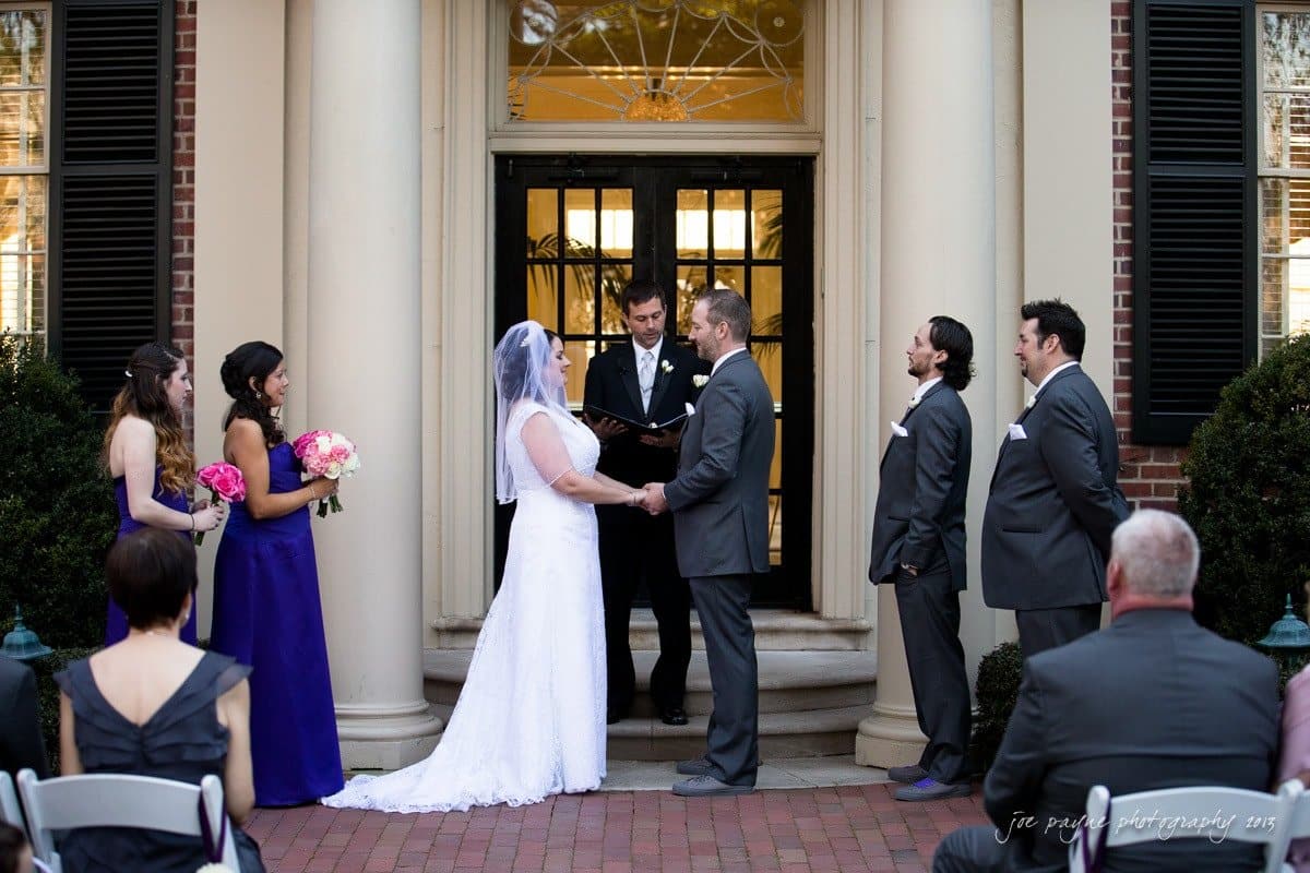 A wedding ceremony in front of a building with columns. The bride in a white dress and the groom in a gray suit hold hands, surrounded by bridesmaids in purple dresses and groomsmen in gray suits. An officiant stands in the doorway.