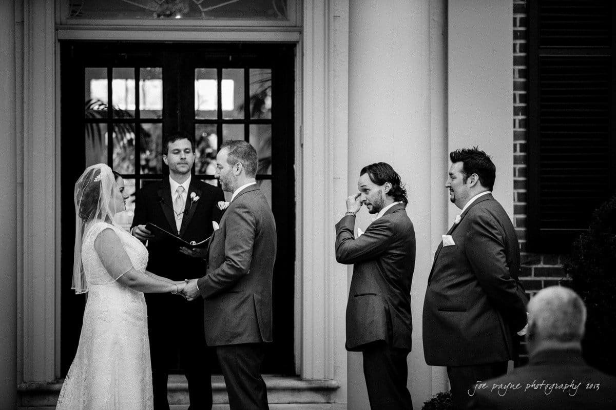 A bride and groom stand holding hands during an outdoor wedding ceremony. The officiant is speaking, while two groomsmen stand nearby. One groomsman wipes his eye. The scene is set in front of a building with large double doors.