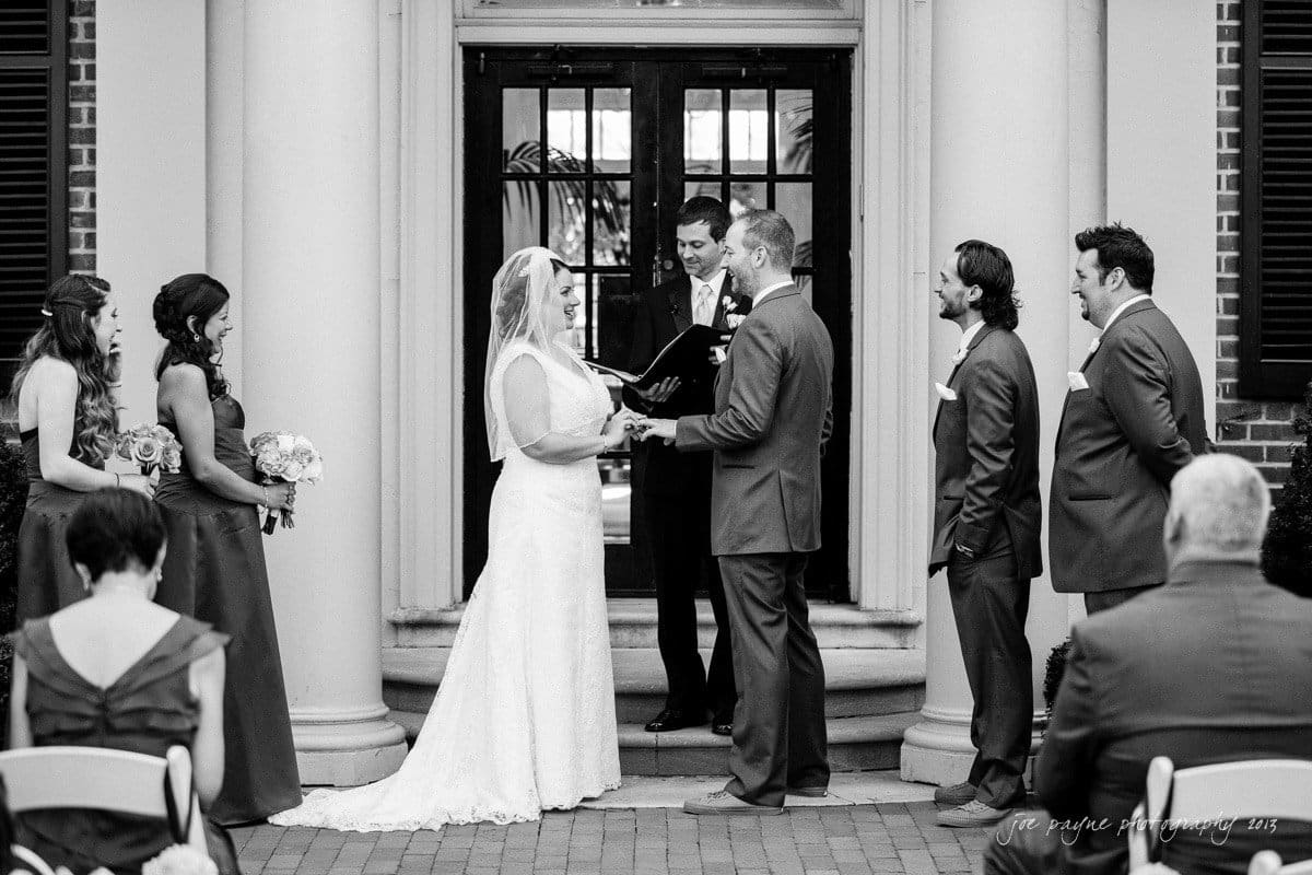 A bride and groom stand at the altar during an outdoor wedding ceremony. The officiant reads from a book, while bridesmaids in dresses and groomsmen in suits stand on either side. Guests are seated watching the ceremony.