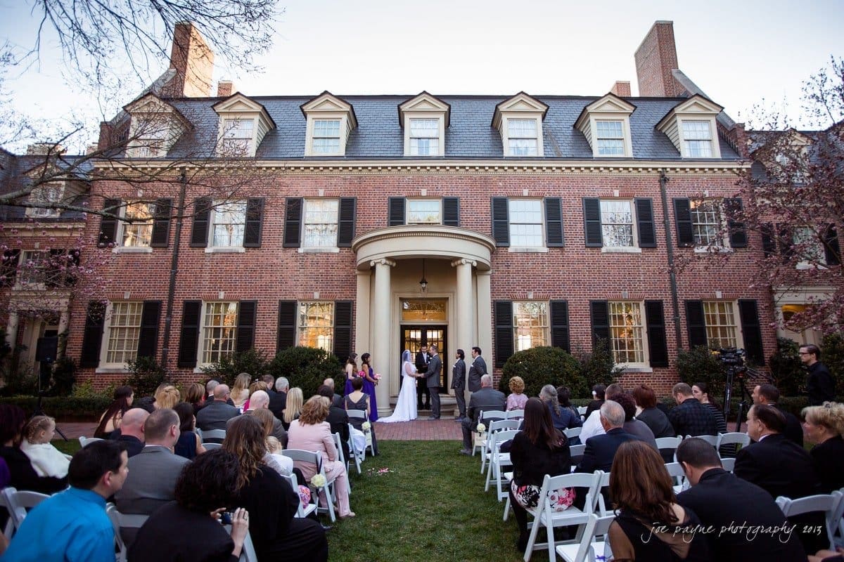 A couple is getting married in an outdoor ceremony, seated guests observe in front of a large, elegant brick building with white trim and black shutters. The scene is surrounded by trees and the sky is clear.