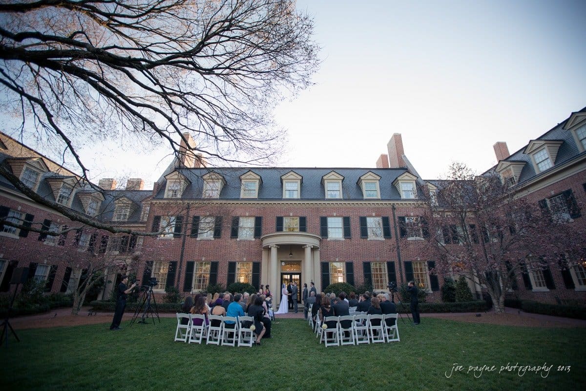A wedding ceremony taking place outdoors in front of a large, elegant building with a brick facade and tall windows. Guests are seated in rows on the lawn, facing the entrance where the ceremony is held. Bare tree branches frame the scene.