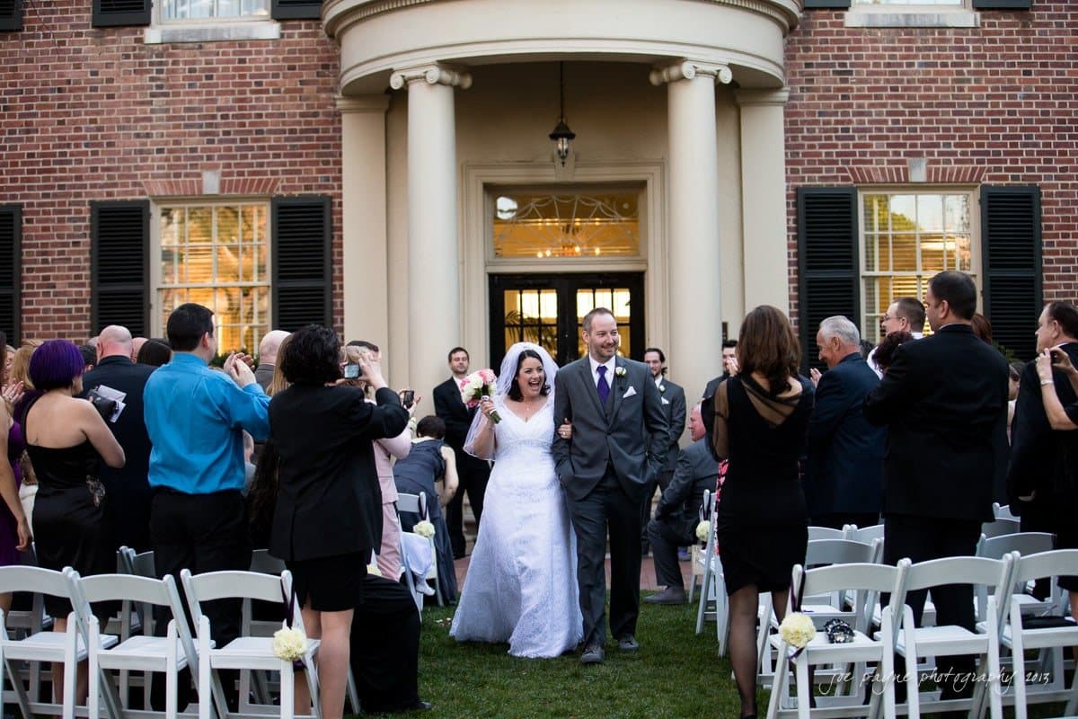 A bride and groom walk down the aisle outdoors, surrounded by guests taking photos. The event takes place in front of a brick building with white columns and black shutters. The couple is smiling and holding hands.