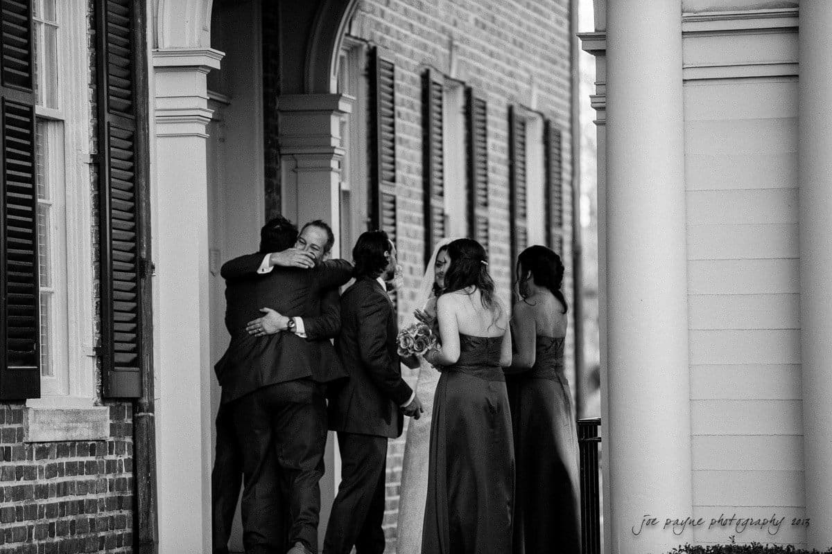 A group of people dressed formally stand on a porch. Two men embrace warmly, while a woman holding a bouquet and another woman walk alongside them. The setting is framed by brick walls and wooden shutters. The image is in black and white.