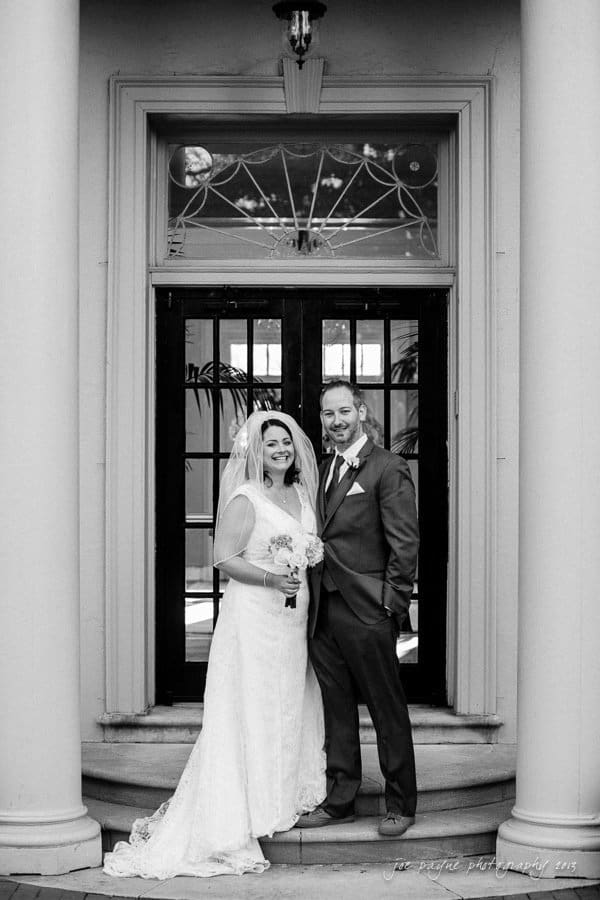 A bride and groom stand smiling in front of a decorative door with glass panels. The bride holds a bouquet and wears a long, lace gown and veil. The groom is dressed in a suit with a boutonniere. They are framed by large, white columns.