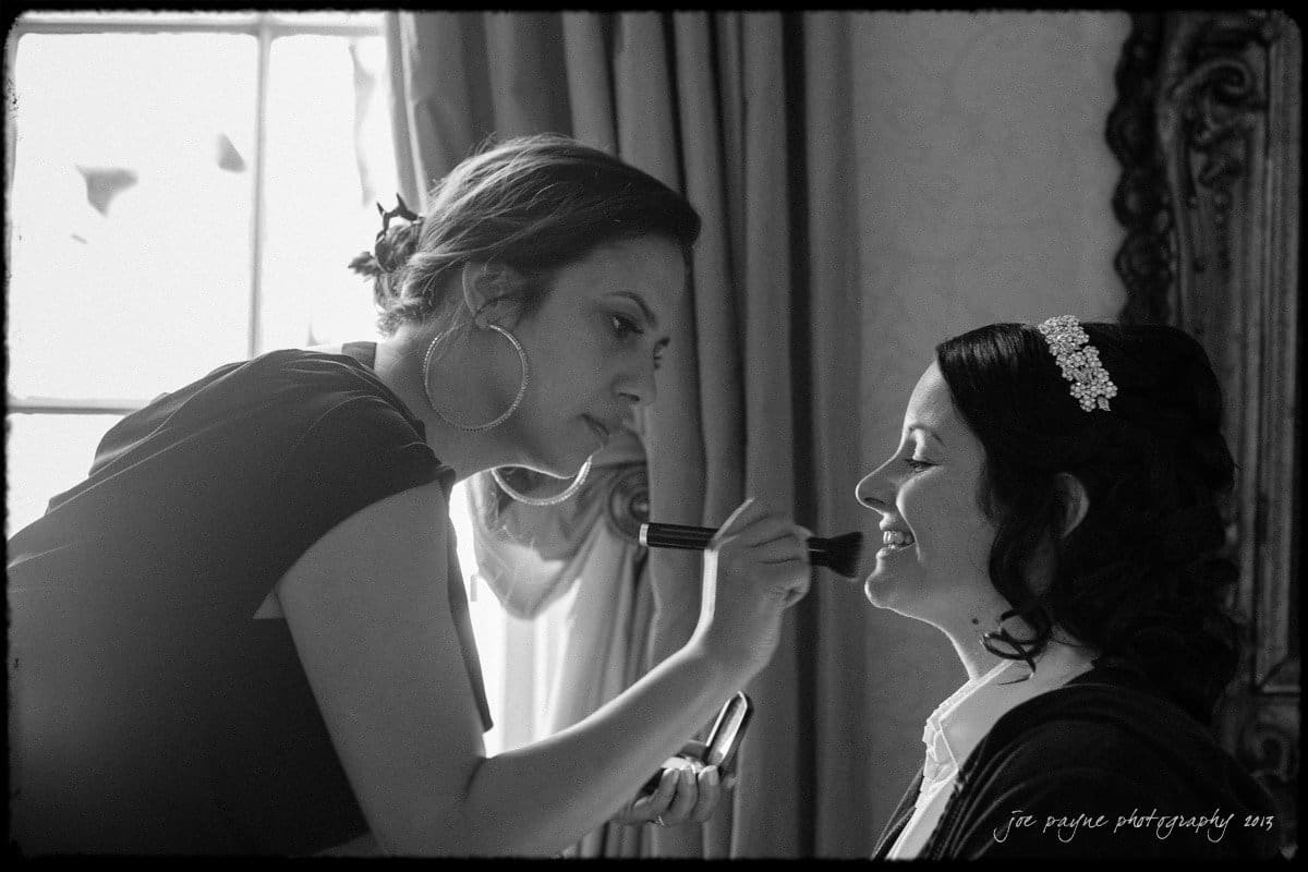 A makeup artist applies makeup to a smiling woman seated near a window. The scene is in black and white, with soft light illuminating the room. The woman wears a decorative headband, and both appear focused on the task.