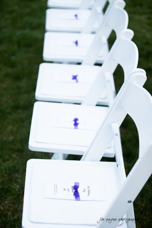 Rows of white folding chairs arranged outdoors on grass, each seat holding a white program tied with a purple ribbon.