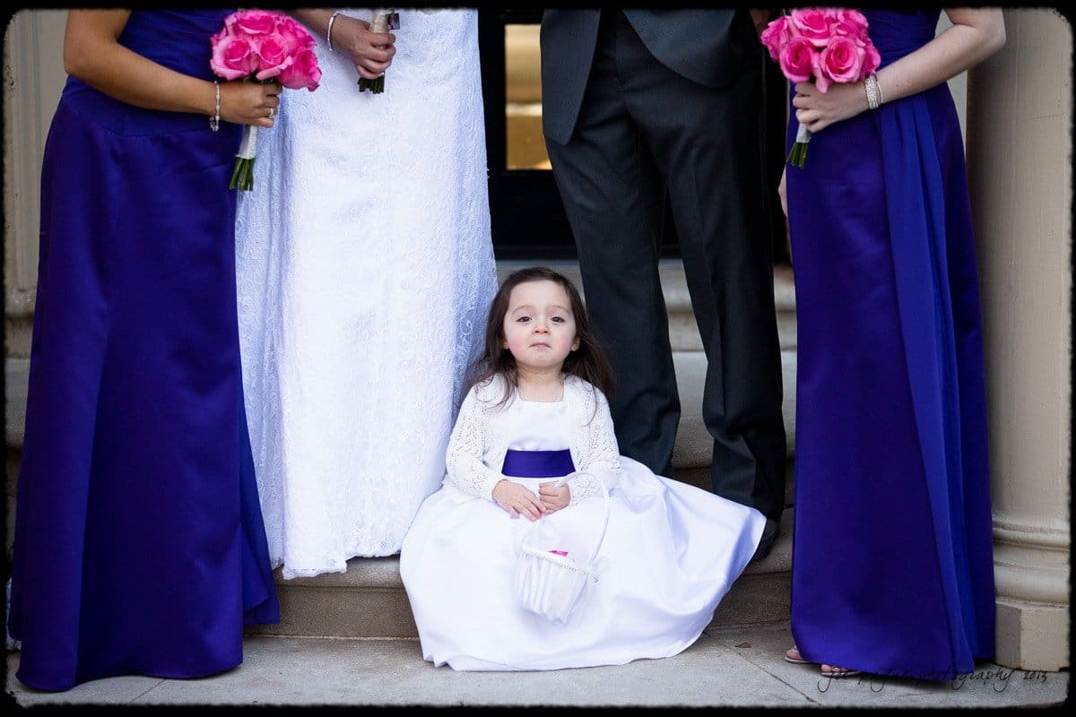 A young girl in a white dress with a purple sash sits on steps, surrounded by adults in formal attire. Three bridesmaids hold pink bouquets, wearing purple dresses, while a man stands in a gray suit.
