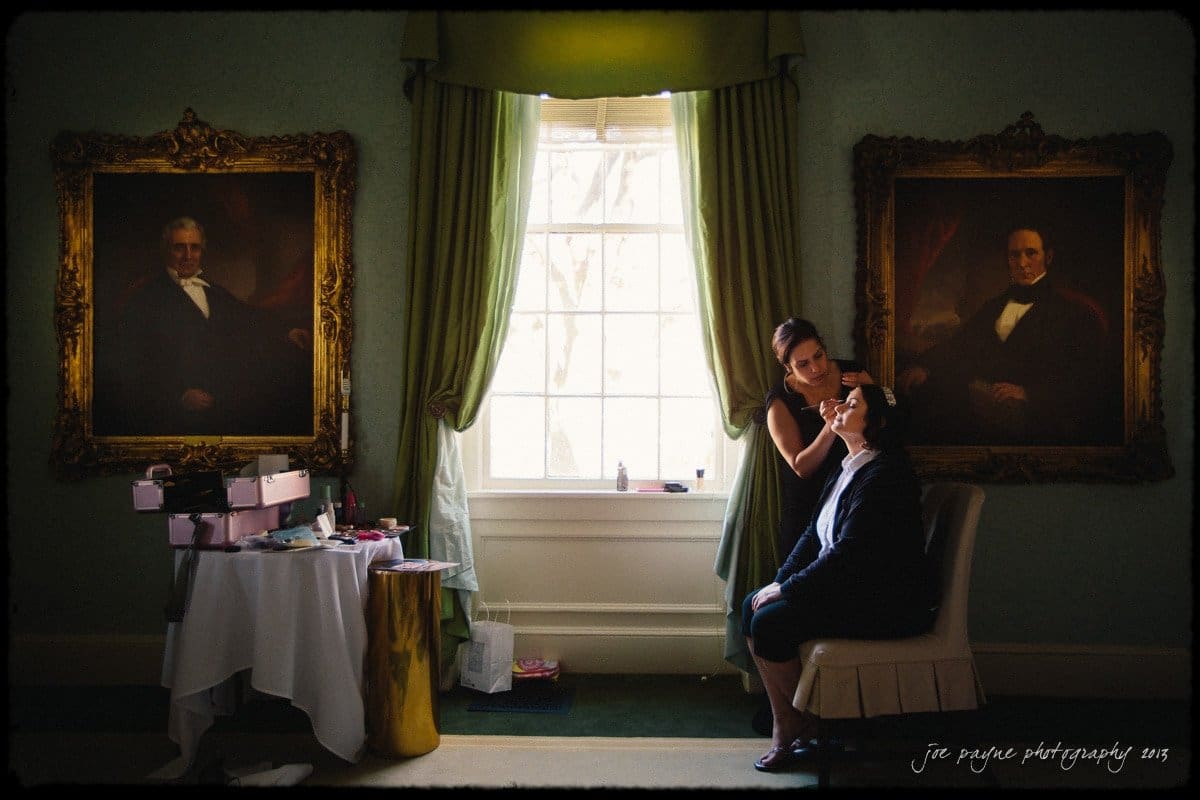 A person is getting makeup applied by another person in a room with ornate green curtains. Two large framed portraits hang on the walls. A table with makeup items is near the window, which provides natural light.
