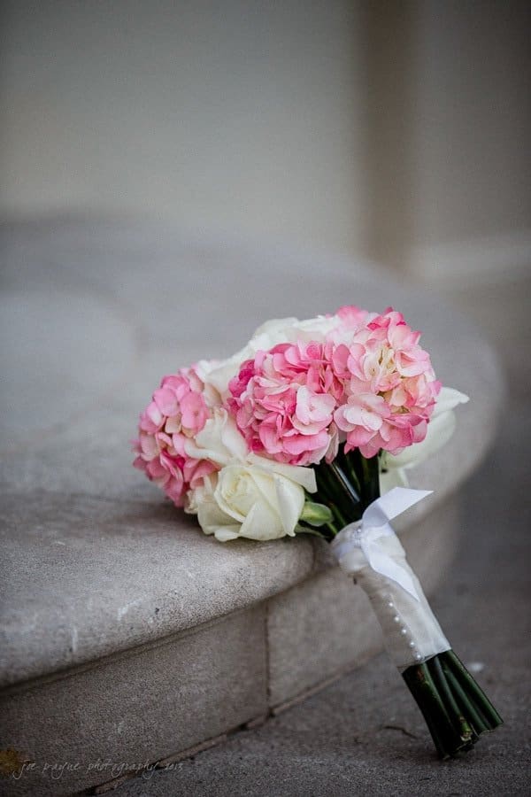 A bouquet of pink hydrangeas and white roses, tied with a white ribbon, rests on stone steps.