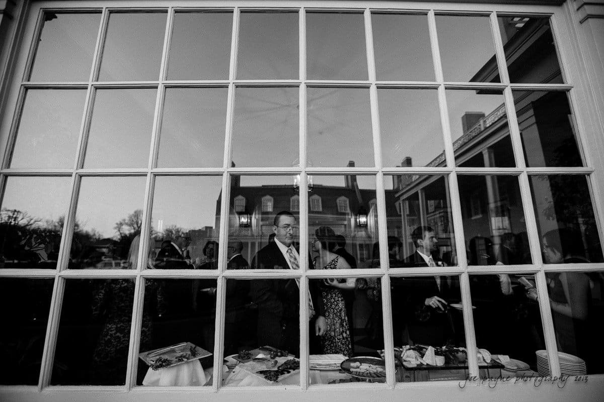 A black and white photo of people inside a building, seen through a large window with multiple panes. Several individuals, dressed formally, are gathered near tables with food. Reflections of the buildings surroundings can be seen on the glass.