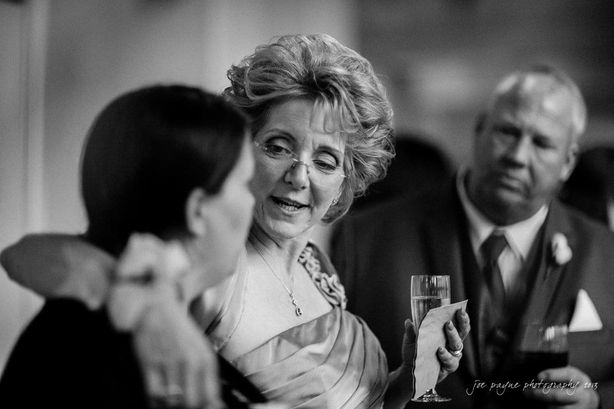 An older woman with glasses, wearing a glamorous dress, holds a champagne glass and talks to a younger person. She has a caring expression. A man in a suit stands nearby, attentively observing the conversation. The photo is in black and white.