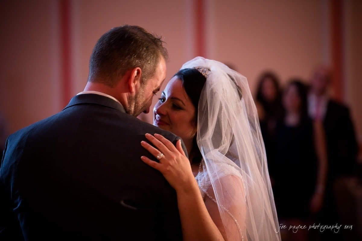 A bride and groom share a close embrace during a dance at their wedding. The bride is smiling softly, wearing a veil, and gently holding the groom. Soft lighting and blurred figures can be seen in the background.