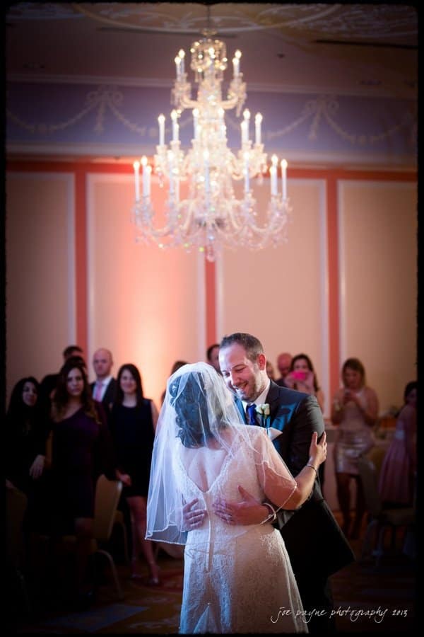 A bride and groom share their first dance under a large, ornate chandelier. The bride wears a veil, and the groom is in a suit. Guests stand in the background, watching the couple with smiles. The setting is an elegant, warmly lit ballroom.