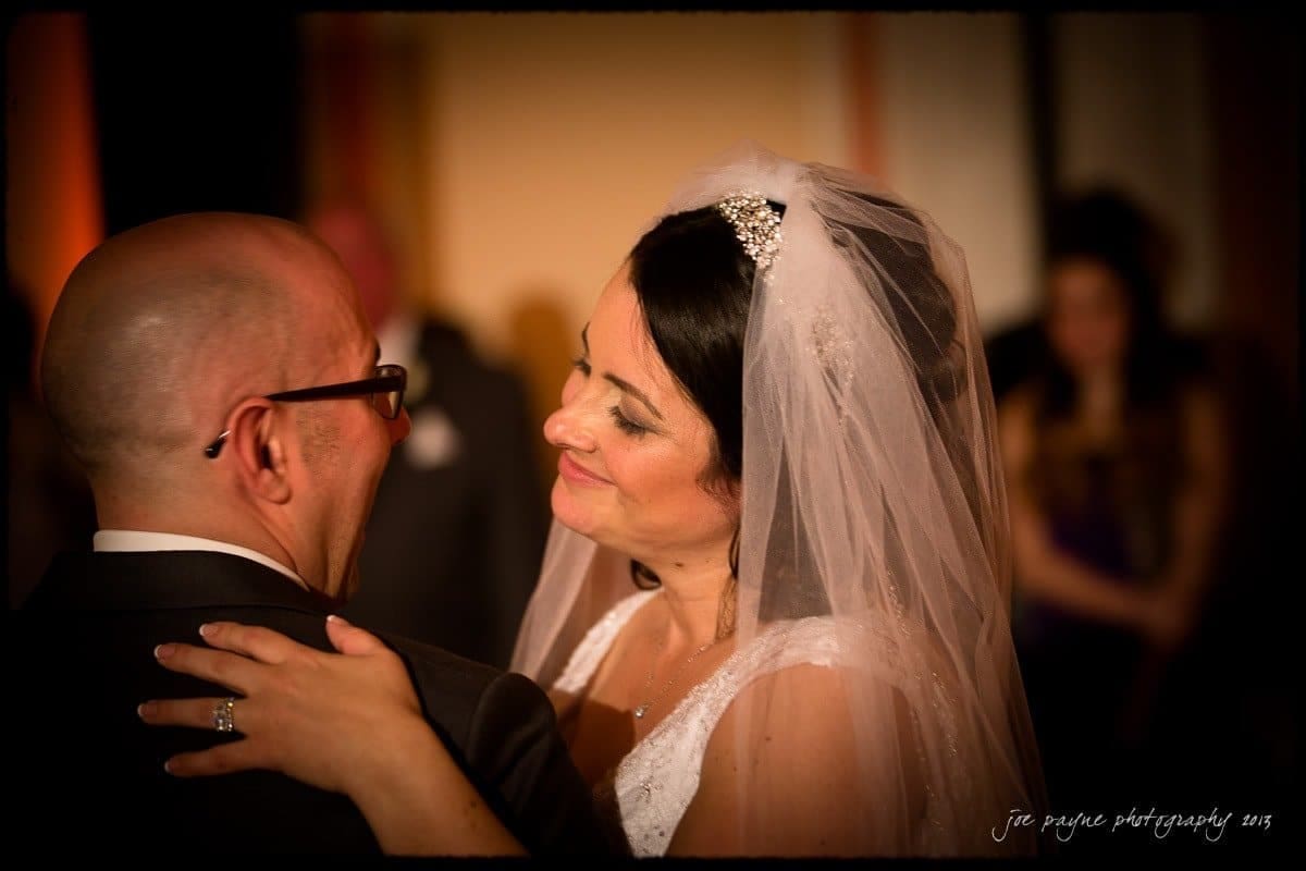 A bride in a white dress and veil smiles at a groom with glasses as they embrace, likely during a wedding dance. The groom is in a suit, and the background is softly blurred with guests.