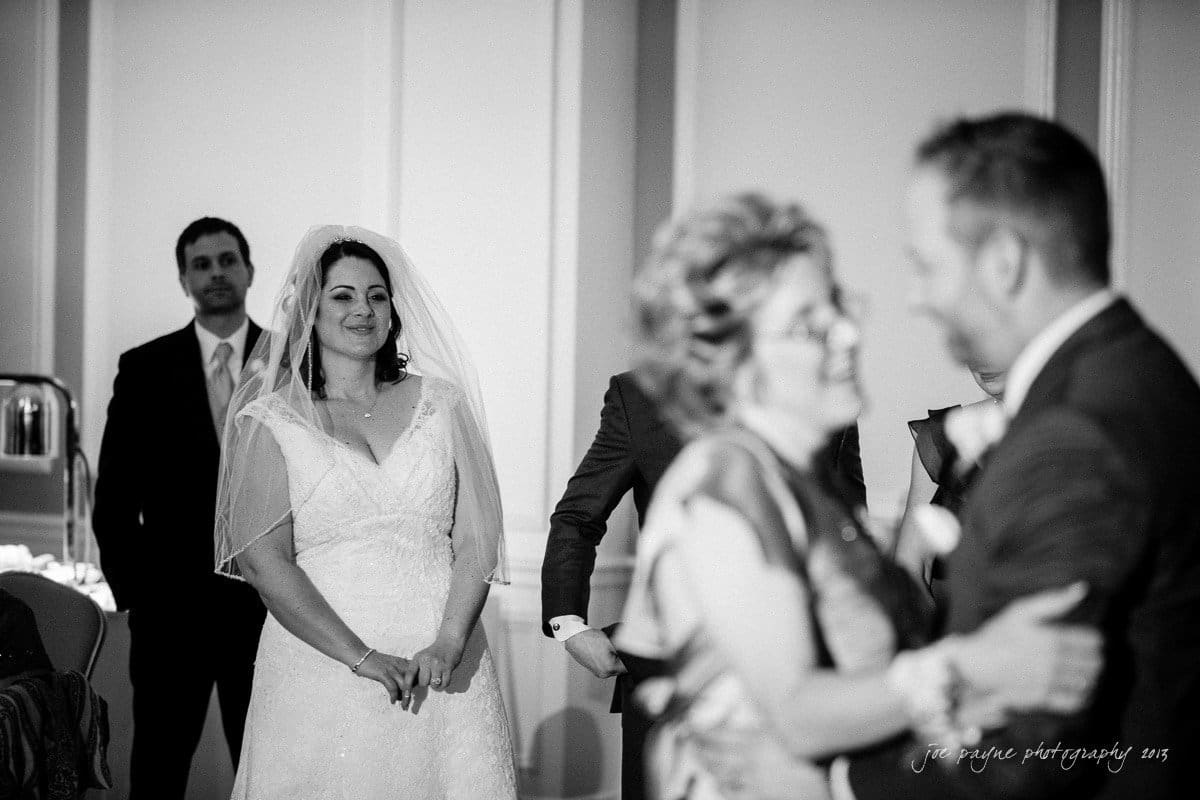 A bride in a wedding dress and veil smiles at a couple dancing closely. A man in a suit stands in the background. The scene is indoors with a formal and joyful atmosphere.