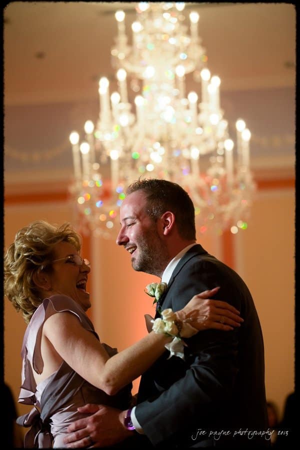 A couple is joyfully dancing in formal attire under a large, elegant chandelier. The woman is wearing a lilac dress, and the man is in a suit with a boutonniere. Both are smiling warmly at each other.