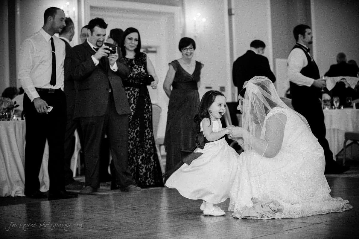 A bride kneels and holds hands with a young girl on the dance floor. Guests in formal attire, some taking photos, watch them with smiles. The room is elegantly decorated for a wedding reception.