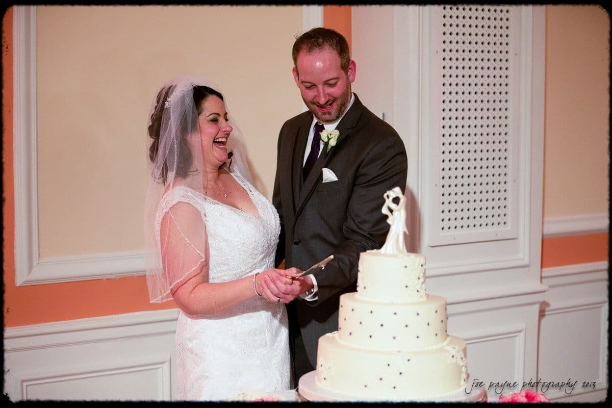 A bride and groom smile as they cut a three-tiered wedding cake. The bride is in a white gown and veil, and the groom is in a dark suit. The cake has a figure topper and small decorations. The setting is an elegantly decorated room.