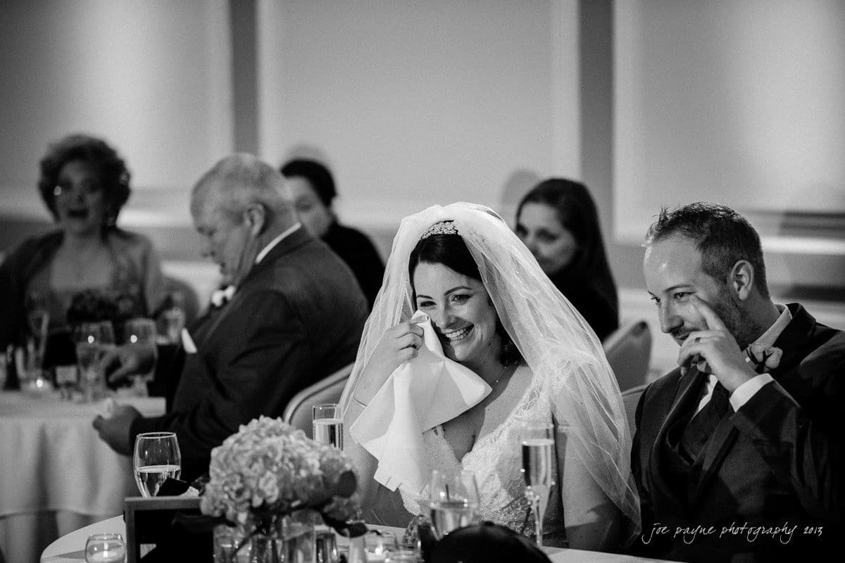 A bride and groom, both in wedding attire, sit at a table laughing. The bride wipes a tear with a napkin. Guests in the background are also smiling, seated at tables with glasses and floral arrangements.