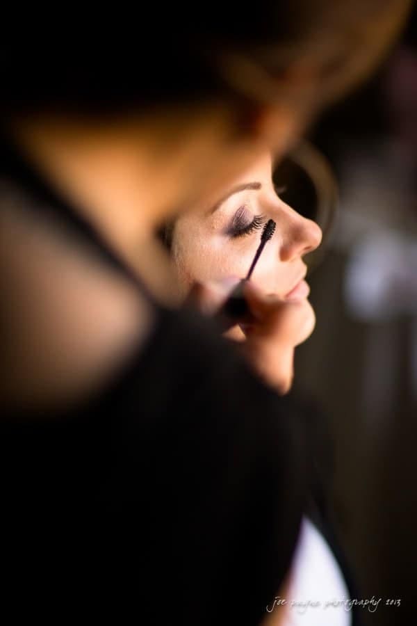 A close-up of a woman having her makeup done. She has her eyes closed while mascara is being applied to her eyelashes. The background is softly blurred, emphasizing her serene expression.