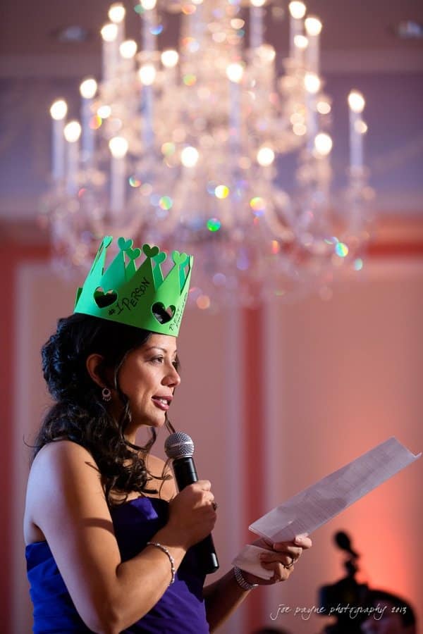 A woman in a strapless dress holds a microphone and a sheet of paper while speaking. She wears a green paper crown with decorations. A sparkling chandelier is visible in the background.