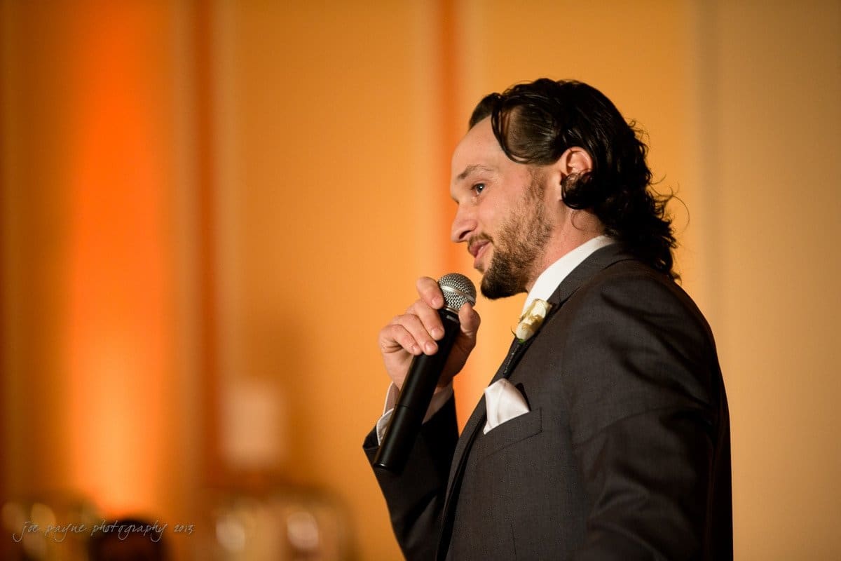 A man in a suit is speaking into a microphone. The background is warmly lit with an orange hue.