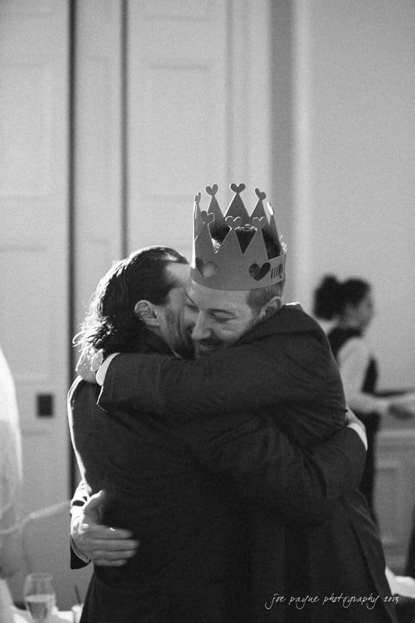Two people in suits share an embrace in a warmly lit room. One person is wearing a paper crown. The background is softly blurred, highlighting the warm connection between them.