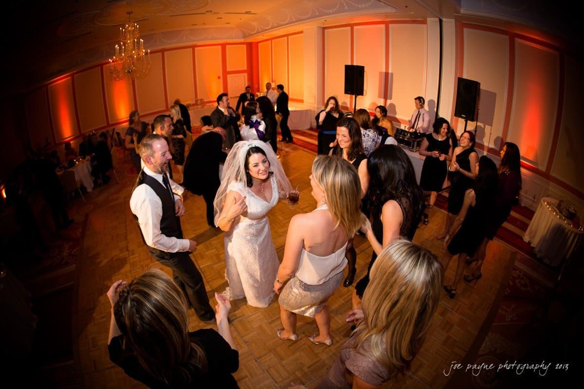 A bride dances joyfully in the center of a circle of friends at a wedding reception. The room is warmly lit with soft orange lights, and guests are dressed in formal attire, enjoying the lively atmosphere.
