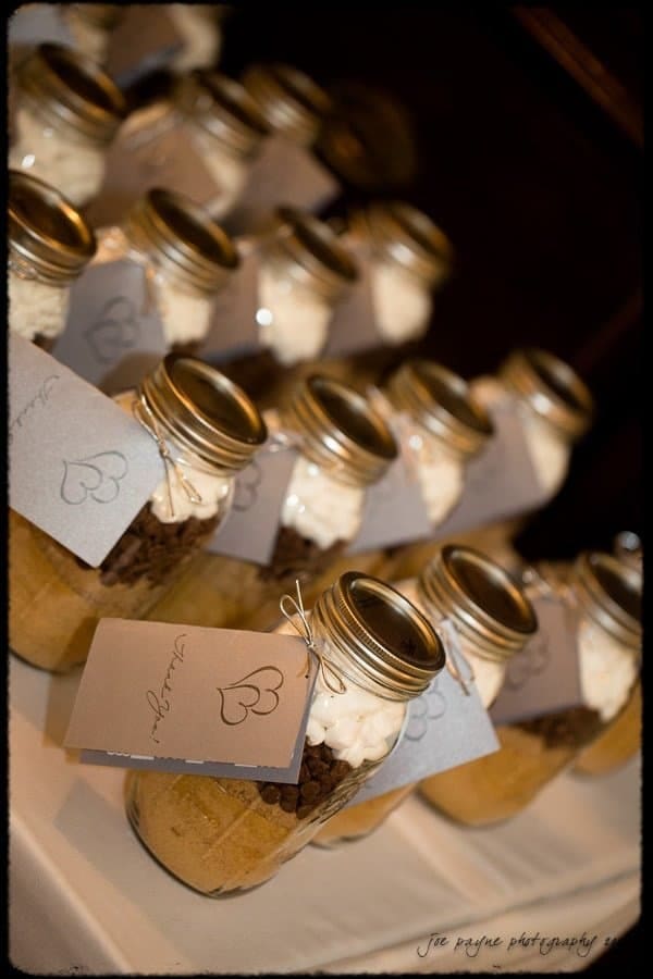 Rows of glass jars filled with layered ingredients, including brown sugar and chocolate chips. Each jar is topped with marshmallows and sealed with a metal lid. Tags with hearts attached to each jar.