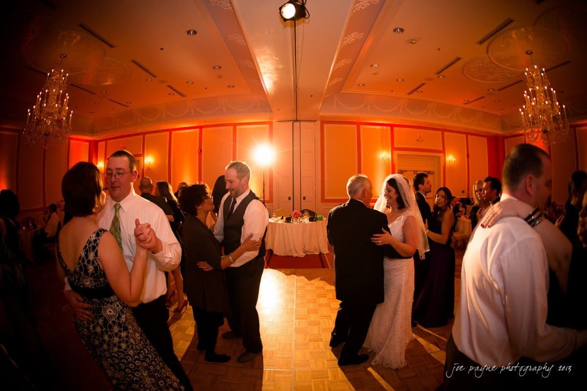 People are dancing in pairs at a warmly lit wedding reception. The bride in a white dress dances with an older man, possibly her father. Other couples are also on the dance floor, surrounded by elegant chandeliers and soft lighting.