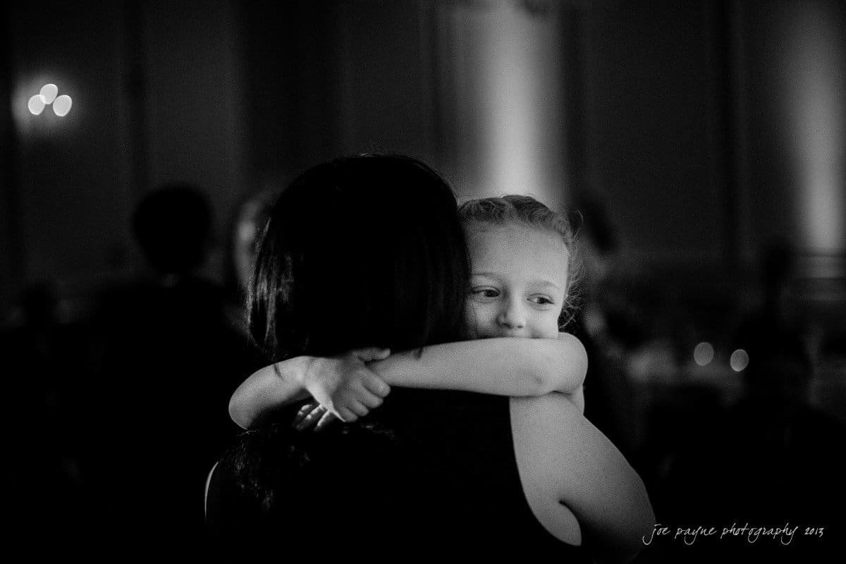 A young girl with a content expression embraces a woman, resting her head on the womans shoulder, in a dimly lit room. The scene conveys warmth and affection.