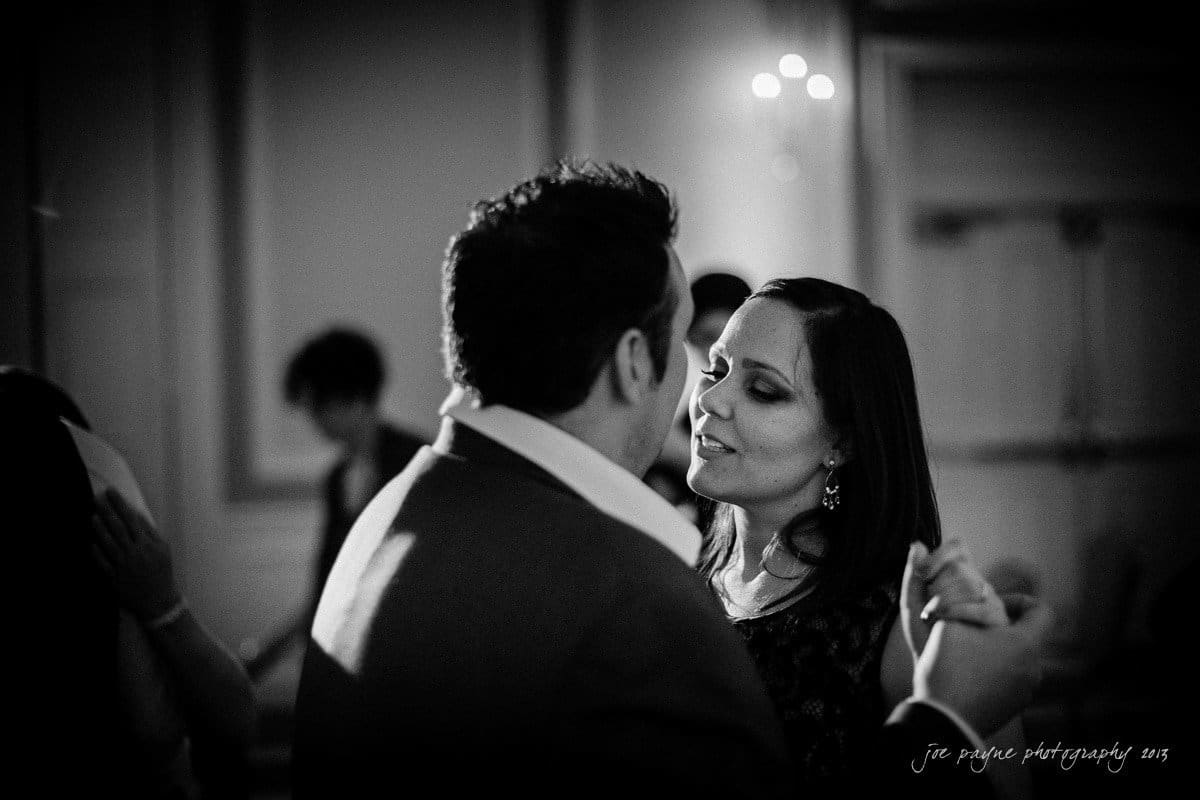 A couple dances closely in a dimly lit room. They are surrounded by blurred figures, creating a soft, intimate atmosphere. The womans eyes are closed as she leans toward the man, who appears to be speaking or singing.