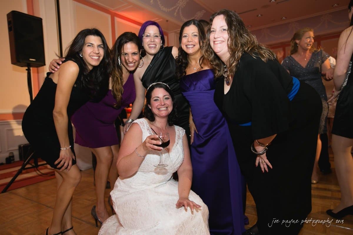 A group of women posing together at a party, smiling for the camera. One woman in a white dress is kneeling in front, holding a glass of wine, while the others stand behind her, dressed in formal attire. The setting is a warmly lit reception hall.