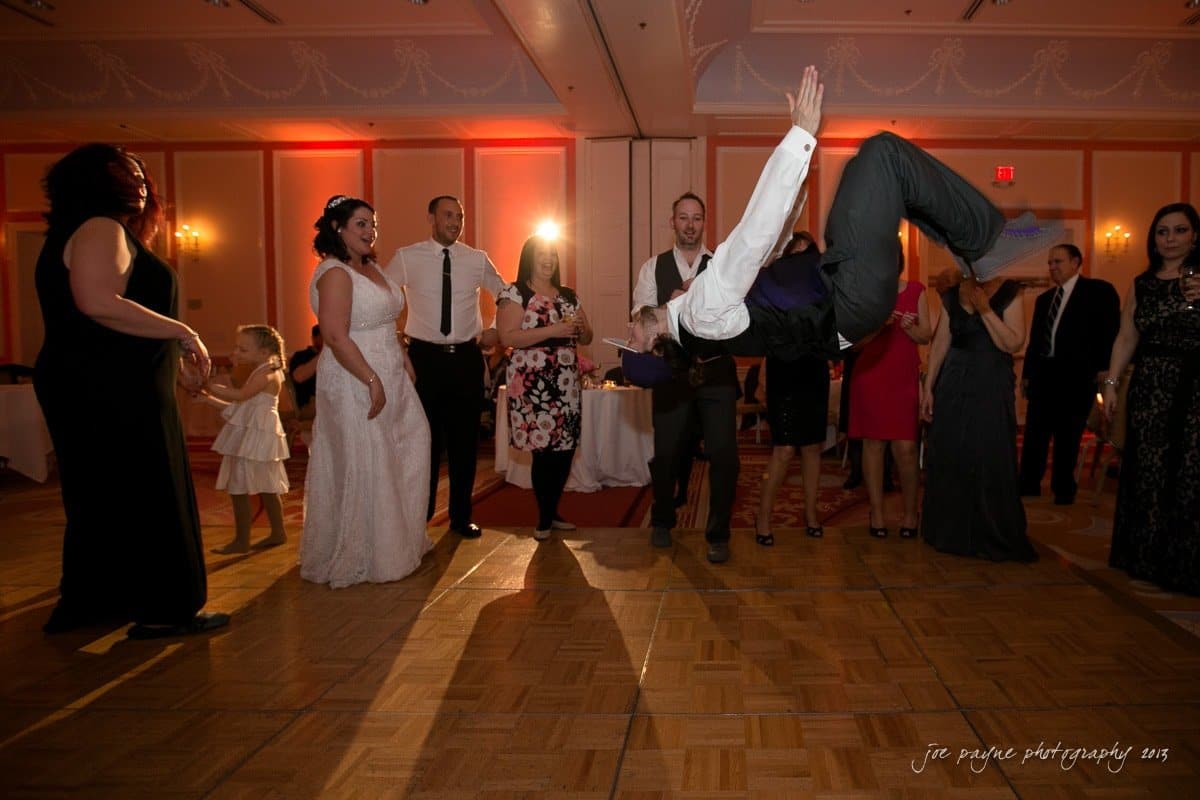 A wedding reception scene with a man performing a backflip on the dance floor. Guests, including a bride in a white dress, watch and react with excitement. The room is dimly lit, creating a festive atmosphere.